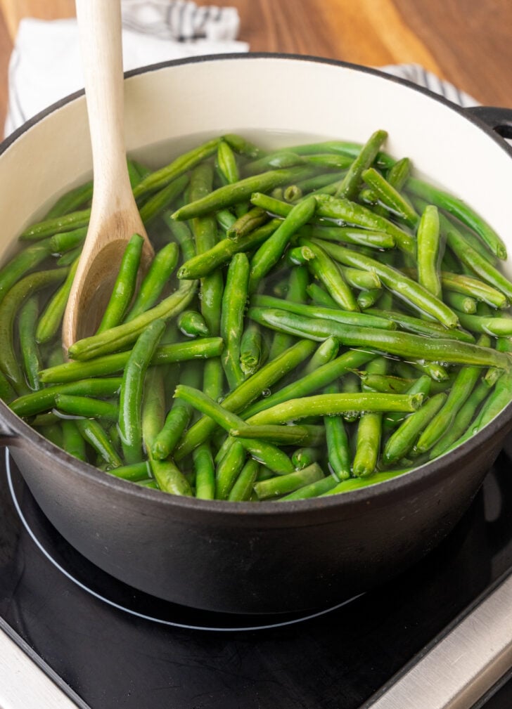 A pot filled with fresh green beans being boiled in water on a stovetop, with a wooden spoon stirring the beans.