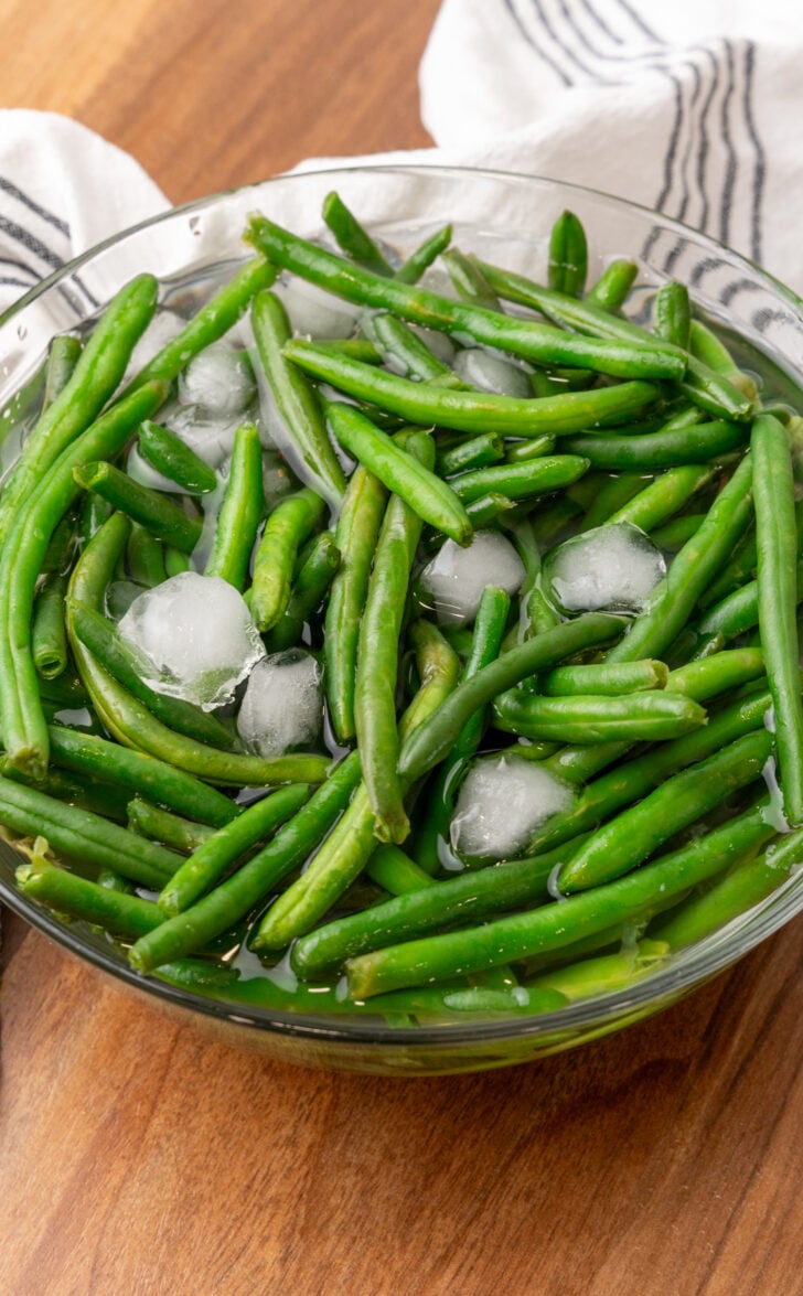 A glass bowl filled with fresh green beans and ice cubes sits on a wooden surface, with a white cloth featuring black stripes in the background.