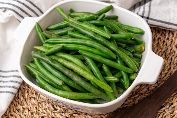 A white dish filled with fresh, cooked green beans sits on a woven placemat, with a striped kitchen towel partially visible in the background.