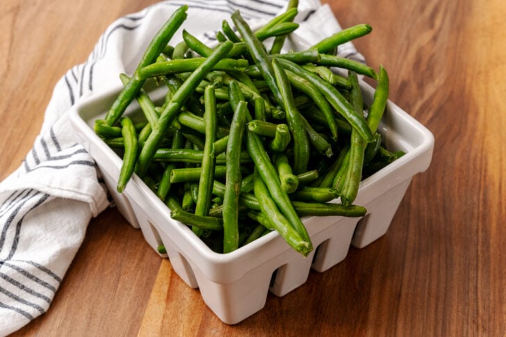 A white ceramic basket filled with fresh green beans sits on a wooden surface next to a white cloth with black stripes.