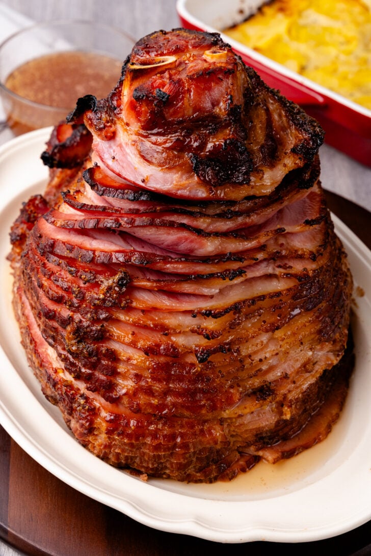 A glazed, spiral-cut ham with caramelized edges is displayed on a white platter. The ham is richly browned and juicy, set on a wooden table. In the background, a dish with a cheesy, layered casserole is partially visible.