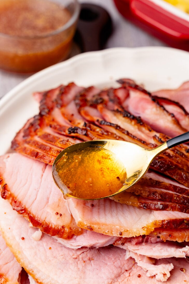 A spoon drizzling glaze over sliced ham on a white plate. In the background, there is a clear glass container with more glaze and a red dish partially visible. The ham has a caramelized crust and pink interior.
