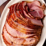 Sliced brown sugar ham 8 on white oval platter with caramelized crust, overhead view on gray wood surface