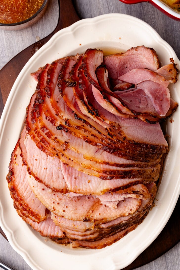 Sliced brown sugar ham 8 on white oval platter with caramelized crust, overhead view on gray wood surface
