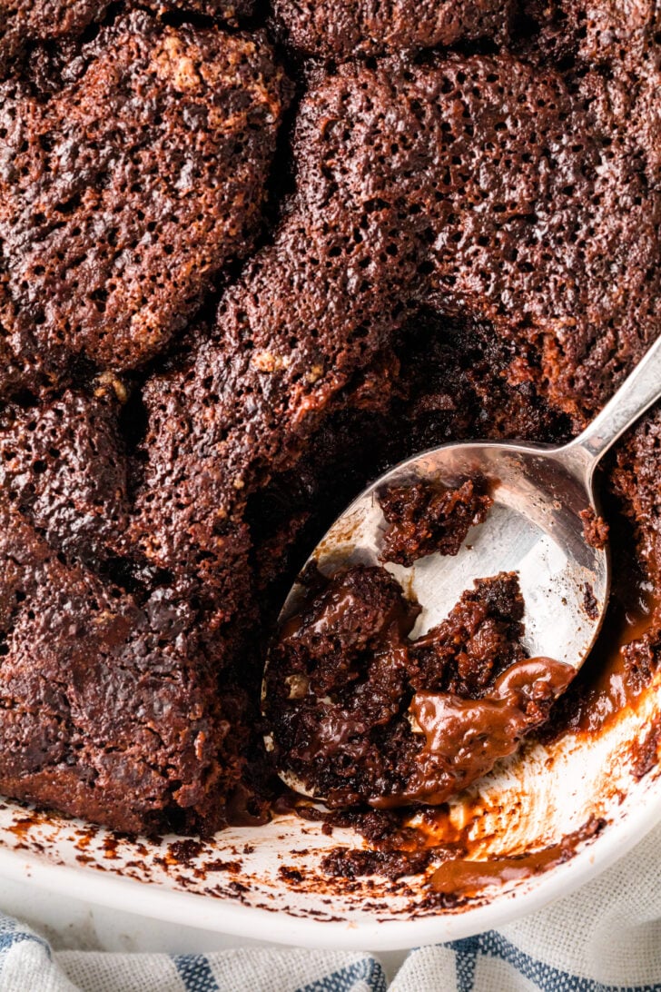 A baking dish filled with rich, gooey chocolate cobbler with a crispy top layer, and a spoon scooping out a warm, molten center of melted chocolate and soft cake.