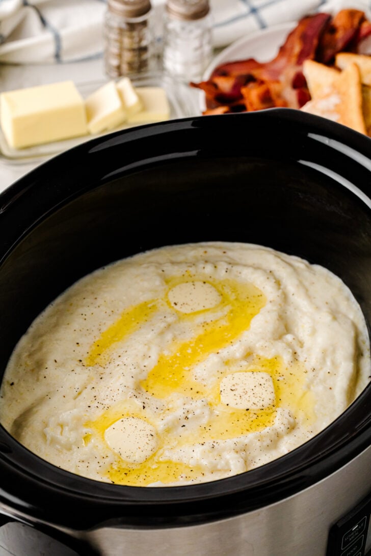 A slow cooker filled with creamy mashed potatoes topped with pats of butter and a sprinkle of pepper; bacon strips, butter slices, and seasonings are visible in the background.
