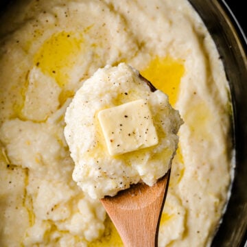 Crock Pot Southern Style Grits in black slow cooker with wooden spoon, topped with butter pat and black pepper