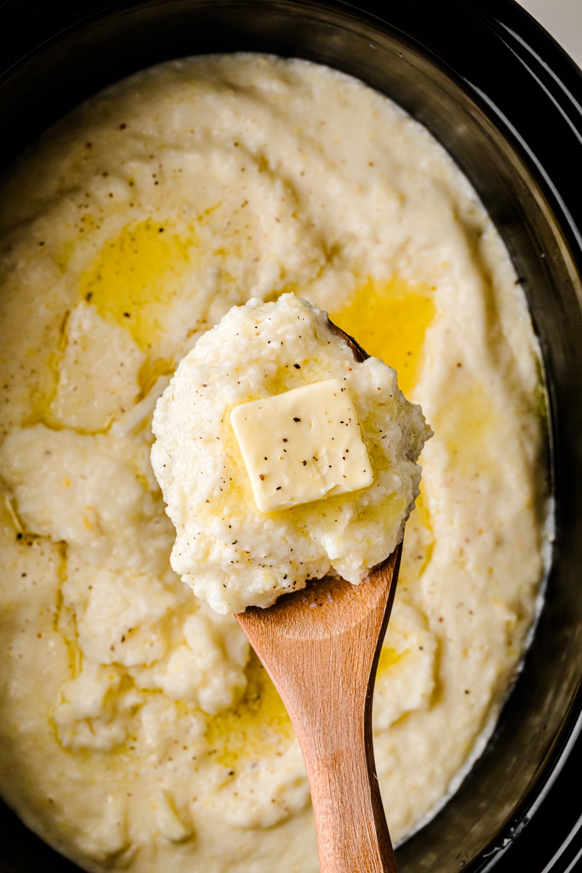 Crock Pot Southern Style Grits in black slow cooker with wooden spoon, topped with butter pat and black pepper