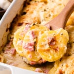 Scalloped Potatoes And Ham in white baking dish with wooden spoon, topped with ham pieces and herbs, close-up overhead view