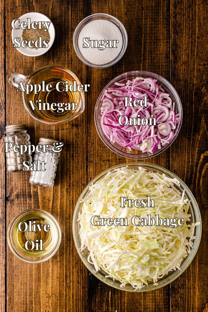 A flat lay of all the vinegar slaw ingredients arranged on a wooden surface, including green cabbage, red onion, apple cider vinegar, olive oil, celery seeds, sugar, and salt and pepper.