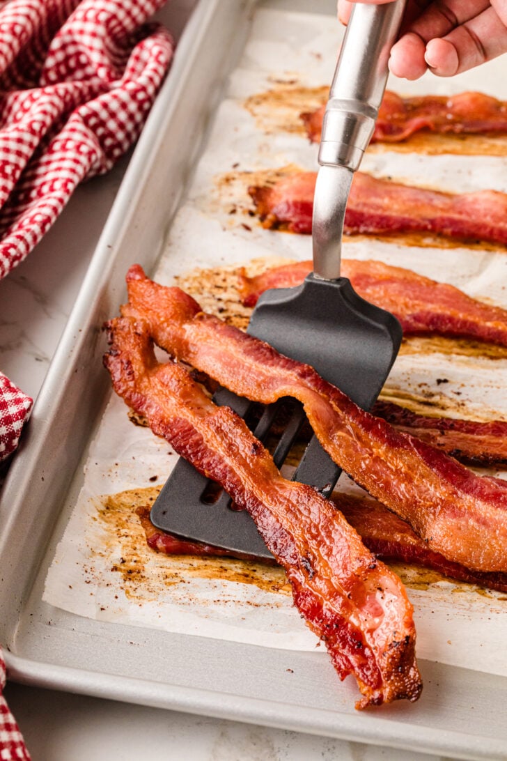 Perfectly cooked bacon scooped up with a spatula from a metal baking pan.