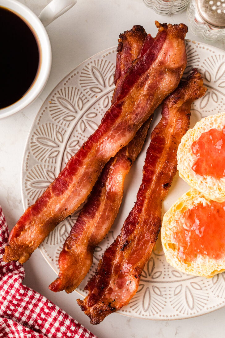 Crispy cooked bacon on a white plate with biscuits topped with jam.