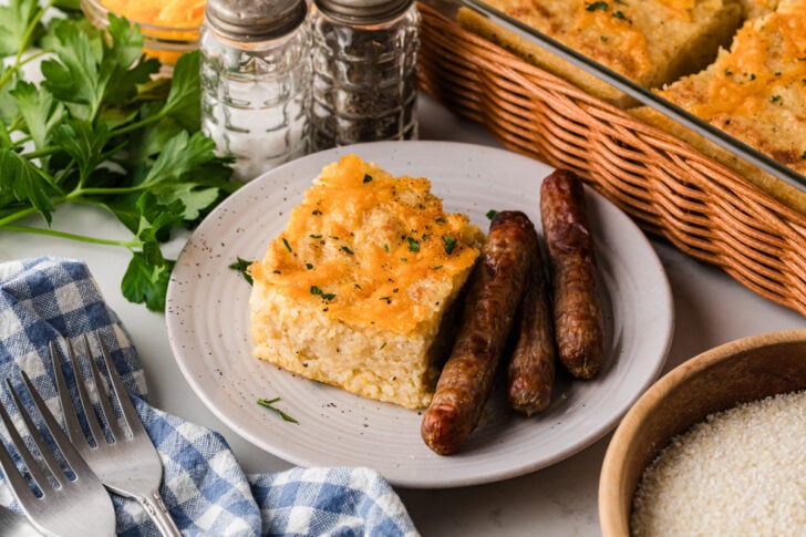 Cheesy cornbread square with crispy sausage links on white plate, garnished with fresh chives and black pepper for Mother's Day Brunch Recip