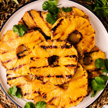 Grilled Pineapple with Brown Sugar Butter Glaze slices on white plate with cilantro garnish and cinnamon sticks on woven placemat