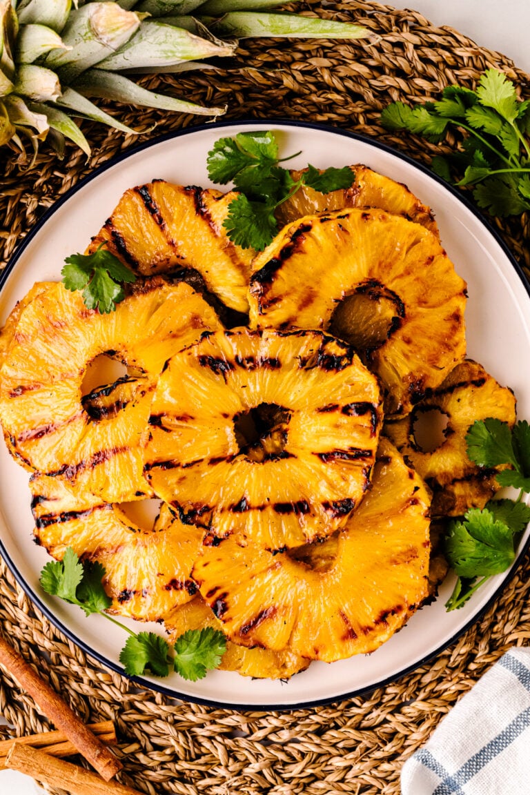 Grilled Pineapple with Brown Sugar Butter Glaze slices on white plate with cilantro garnish and cinnamon sticks on woven placemat