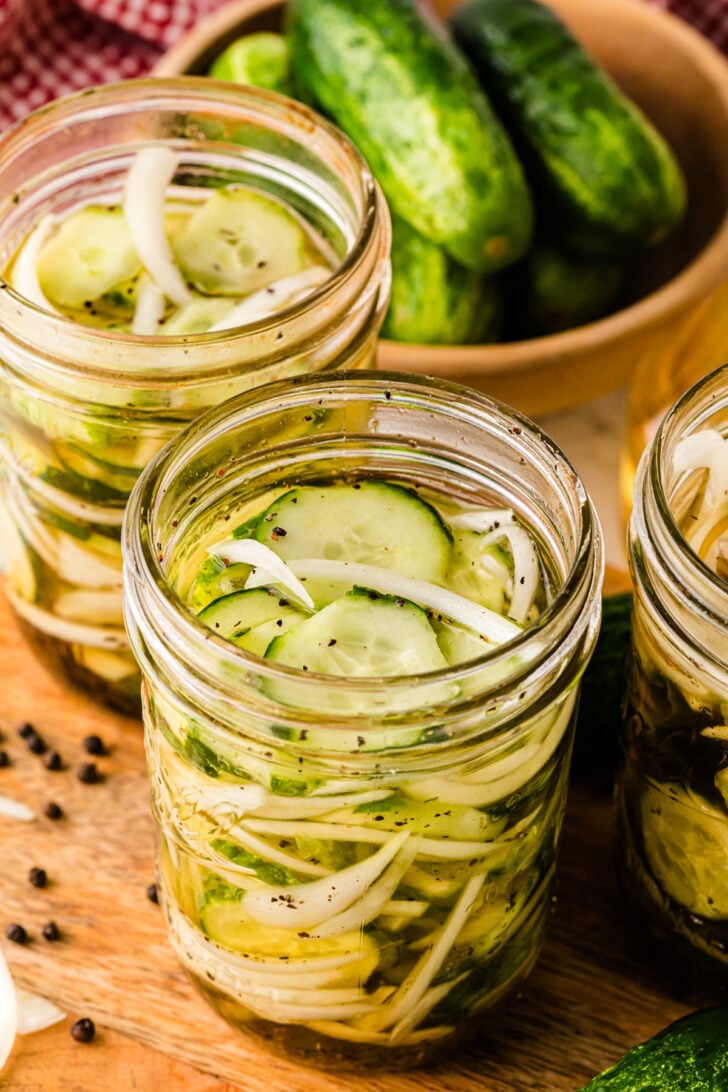 Glass mason jars filled with homemade cucumber and onion pickles in a vinegar brine, sitting on a wooden surface. Whole cucumbers in a bowl and scattered peppercorns are visible in the background, adding a rustic touch.