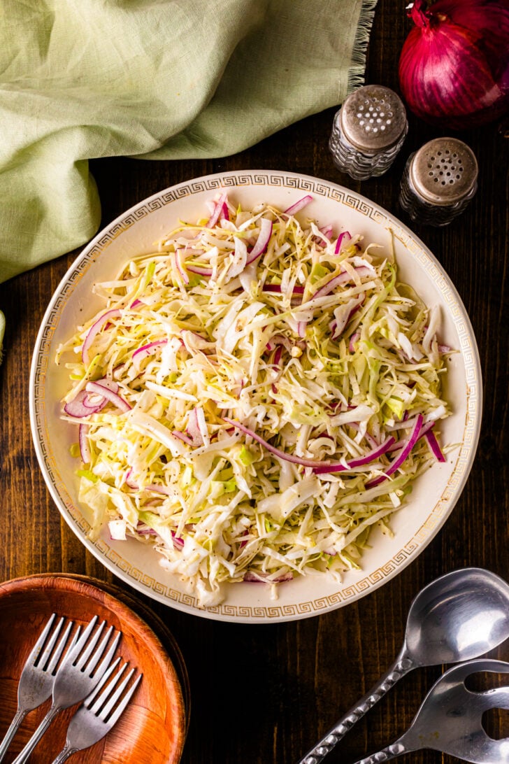 Overhead view of a wooden salad bowl filled with vinegar slaw, featuring shredded green cabbage and red onion, with black pepper sprinkled on top.