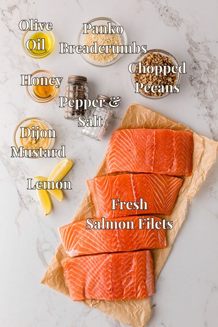 A top-down view of a marble kitchen counter with ingredients for a salmon dish arranged around a parchment-lined plate holding three fresh salmon fillets. The ingredients include a bowl of olive oil, a jar of honey, a bowl of Dijon mustard, a lemon cut into slices, a pepper grinder and salt shaker, a bowl of panko breadcrumbs, and a bowl of chopped pecans, each labeled with its name.