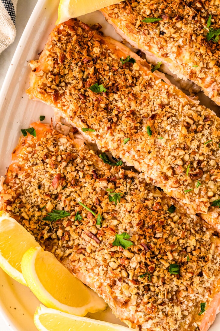 A close-up view of a white platter holding three pieces of baked salmon, each topped with a generous layer of chopped nuts and garnished with fresh parsley. Slices of lemon are arranged around the salmon, and a checkered cloth is partially visible in the background.