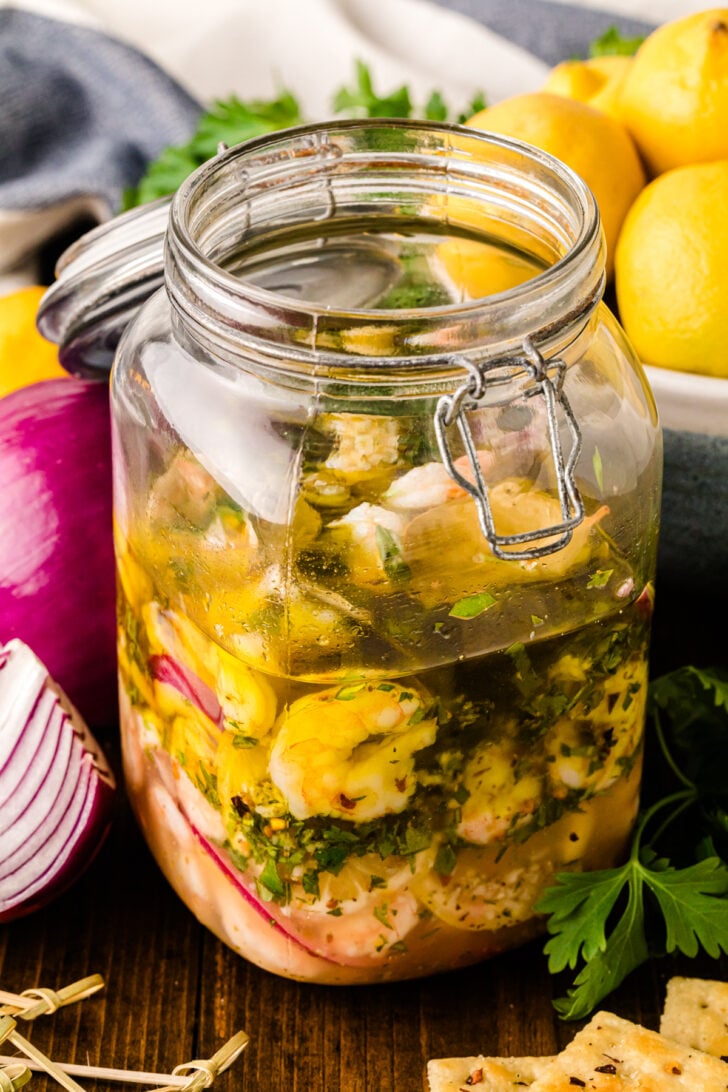 A glass jar filled with pickled shrimp, red onion slices, herbs, and lemon slices in a tangy brine. The jar is surrounded by fresh lemons, red onion, crackers, and parsley on a wooden surface.