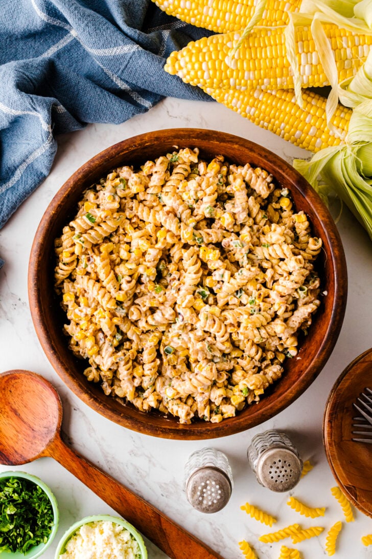 Overhead view of creamy street corn pasta salad in a large wooden bowl, surrounded by fresh corn on the cob, salt and pepper shakers, a wooden spoon, and small bowls of chopped parsley and crumbled cheese.