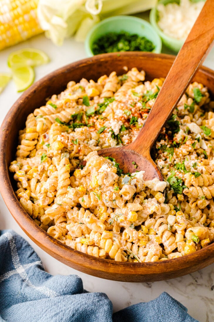 A wooden bowl filled with creamy Street Corn Pasta Salad made with rotini pasta, charred corn, crumbled cheese, chopped herbs, and spices. A wooden spoon is resting in the bowl, ready to serve. Lime wedges and fresh ingredients are blurred in the background on a marble surface.