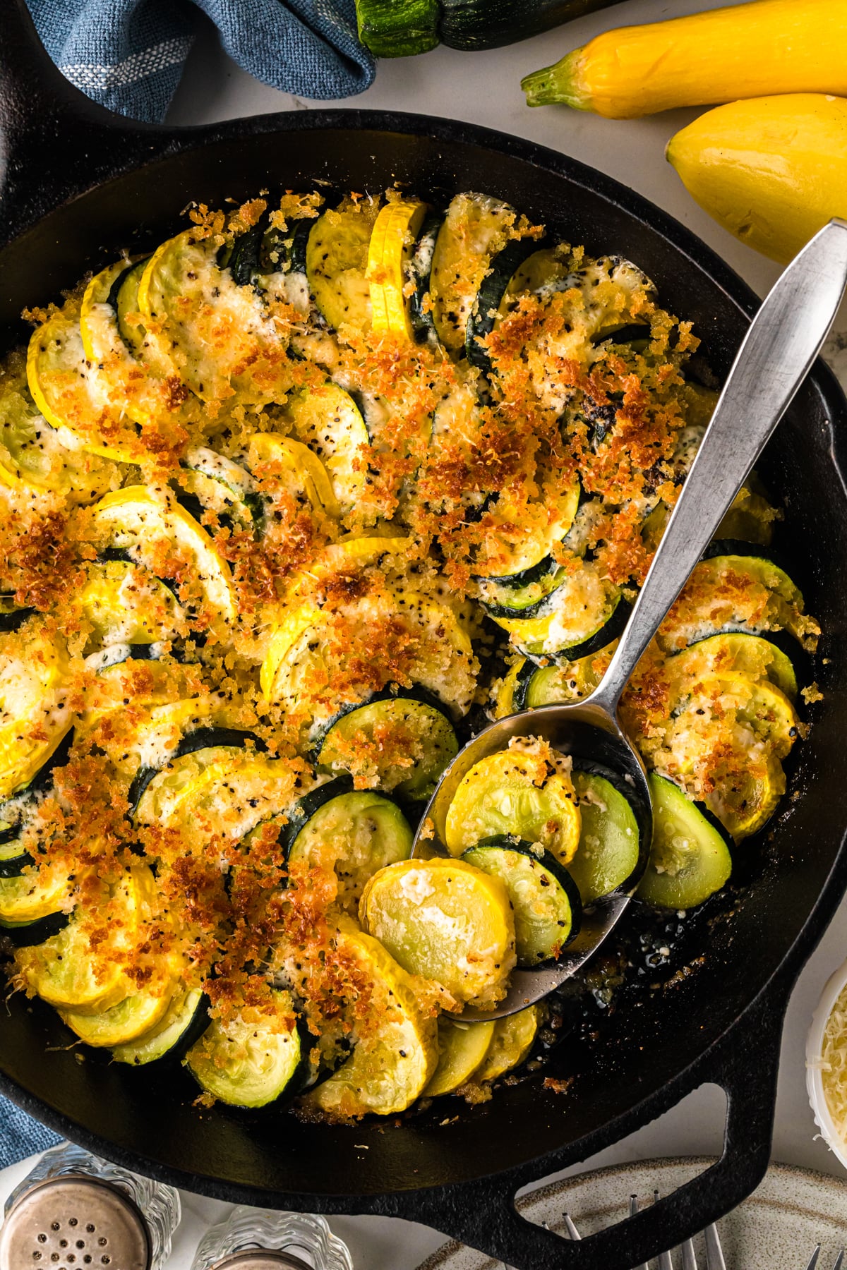 Golden-brown zucch squash casserole 8 in cast iron skillet with crispy breadcrumb topping and fresh yellow squash beside it