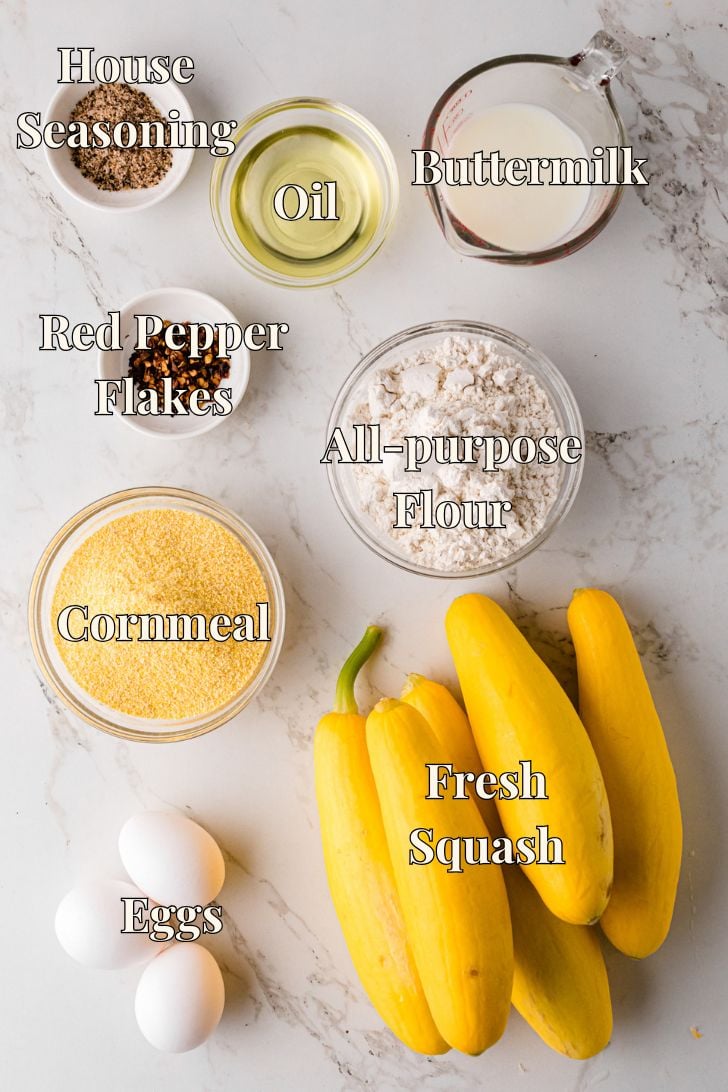 Flat lay of labeled ingredients for fried squash on a marble surface, including fresh yellow squash, eggs, cornmeal, all-purpose flour, red pepper flakes, house seasoning, oil, and buttermilk, each in bowls or measuring cups.
