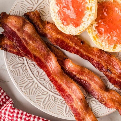 Crispy bacon strips and grapefruit halves with jam on a decorative plate beside black coffee and a red checkered napkin