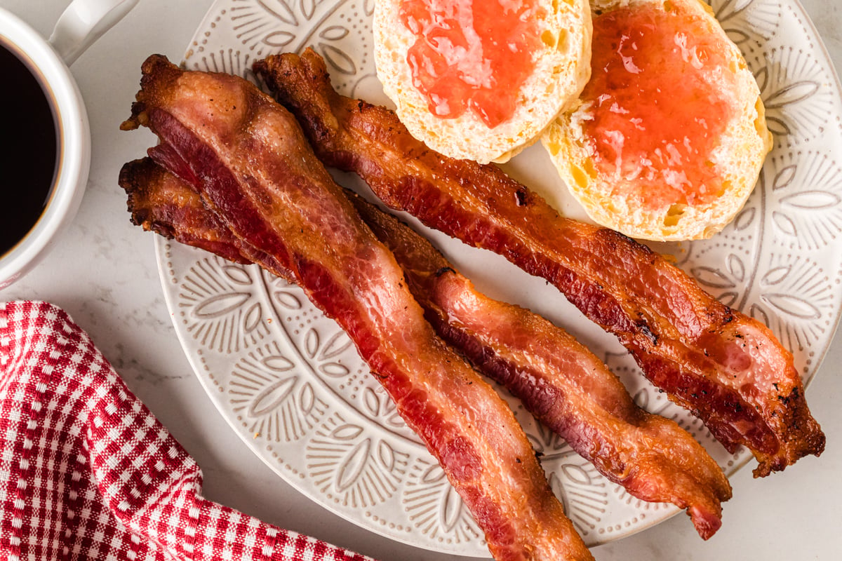 Crispy bacon strips and grapefruit halves with jam on a decorative plate beside black coffee and a red checkered napkin