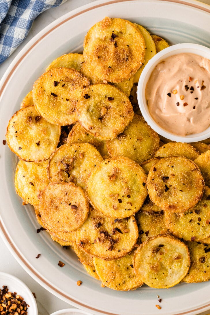 A plate of crispy, golden-brown fried squash rounds sprinkled with red pepper flakes, served with a bowl of creamy dipping sauce. The dish sits on a light plate with a checkered cloth in the background.