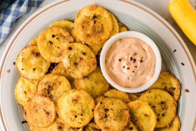 Plate of golden fried squash with crispy edges and black pepper seasoning, served with creamy tan dipping sauce on white dish