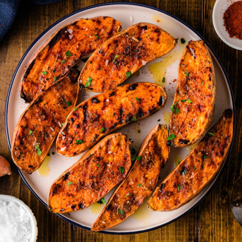 Grilled sweet potatoes 5 arranged on white plate with paprika seasoning, fresh parsley garnish, and olive oil drizzle on wooden table