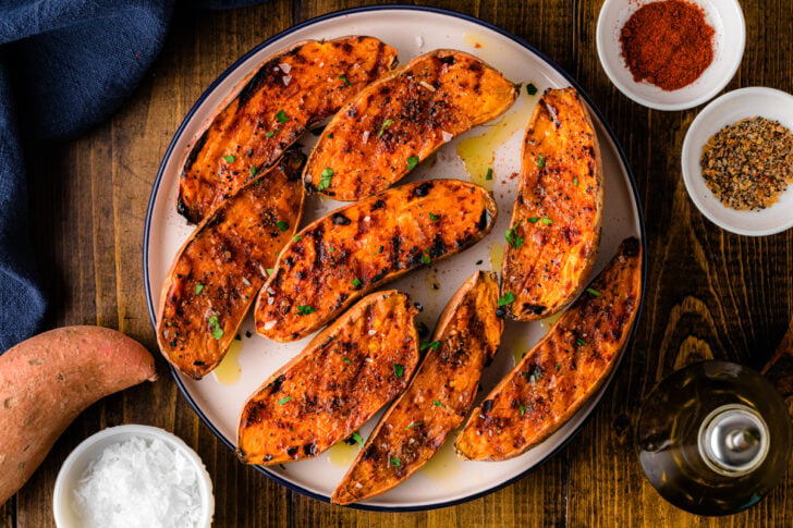 Grilled sweet potatoes 5 arranged on white plate with paprika seasoning, fresh parsley garnish, and olive oil drizzle on wooden table