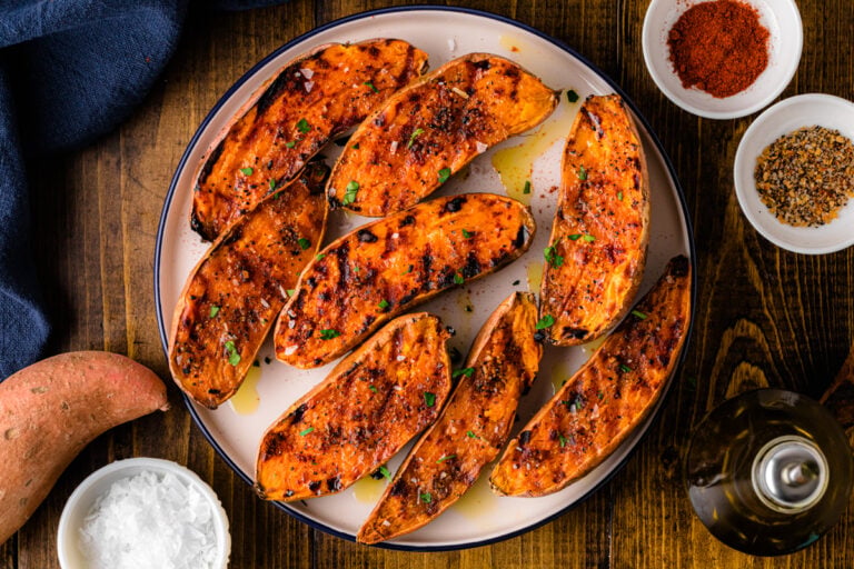 Grilled sweet potatoes 5 arranged on white plate with paprika seasoning, fresh parsley garnish, and olive oil drizzle on wooden table