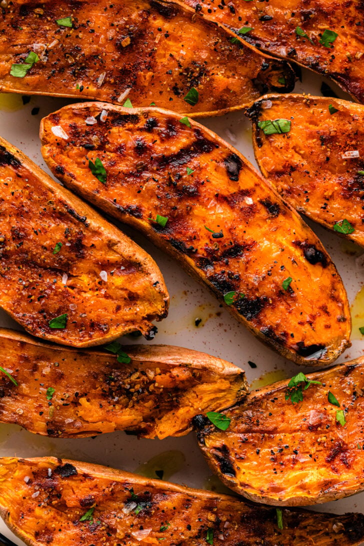 Close-up of grilled sweet potato halves with charred grill marks, sprinkled with flaky sea salt, cracked pepper, and fresh parsley. The potatoes have a caramelized, golden-orange surface with crispy edges.