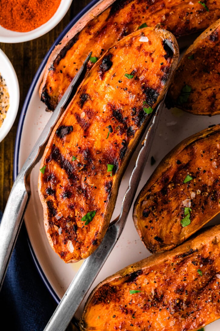 Grilled sweet potato half being lifted with metal tongs, showing deep grill marks, charred edges, and sprinkled sea salt and herbs. Surrounding sweet potatoes are arranged on a white plate with bowls of spices nearby.