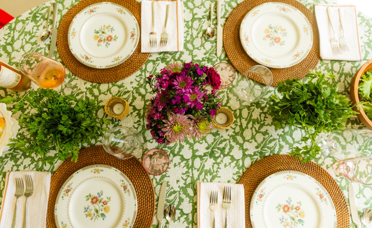 Overhead table setting for four with floral plates on woven chargers, green botanical tablecloth, magenta daisies, and glassware for 20+ Rec