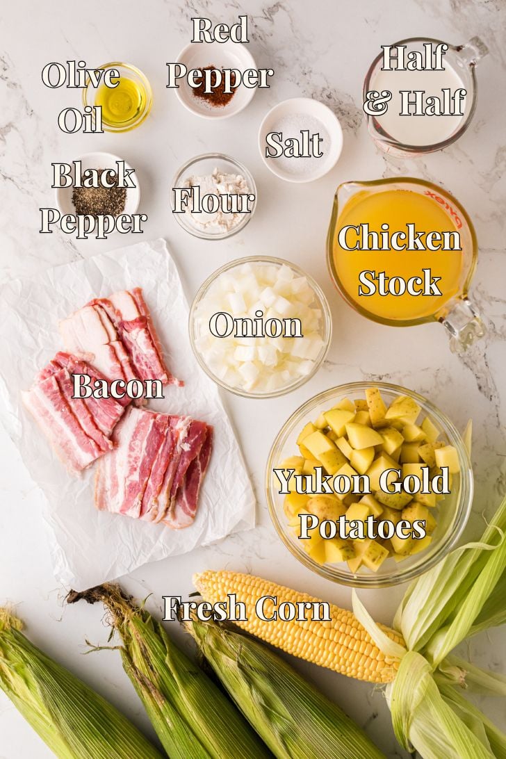 Overhead flat lay of corn chowder ingredients on a marble surface: fresh corn on the cob, bacon strips, diced Yukon gold potatoes, diced onion, chicken stock, half-and-half, flour, olive oil, black pepper, red pepper, and salt—each labeled.