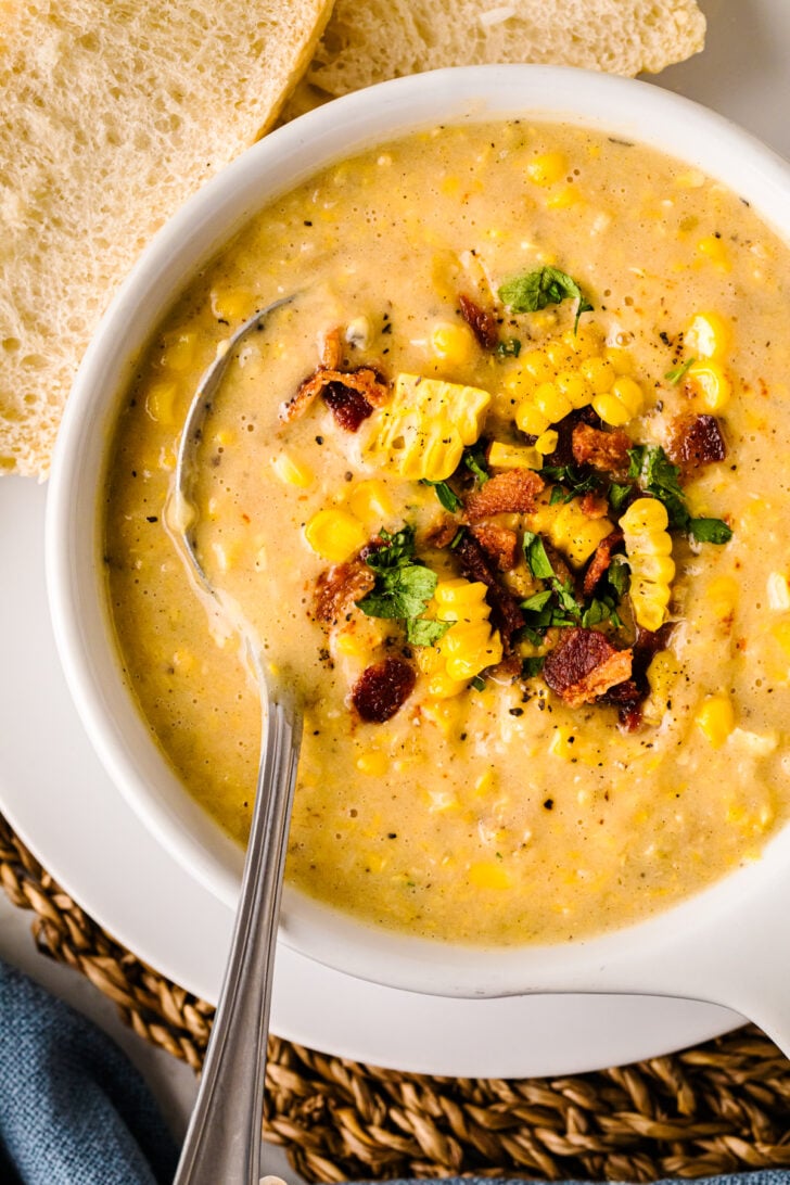Overhead bowl of creamy corn chowder topped with sweet corn kernels, crispy bacon, and parsley, with a spoon in the bowl and sliced bread on the side.