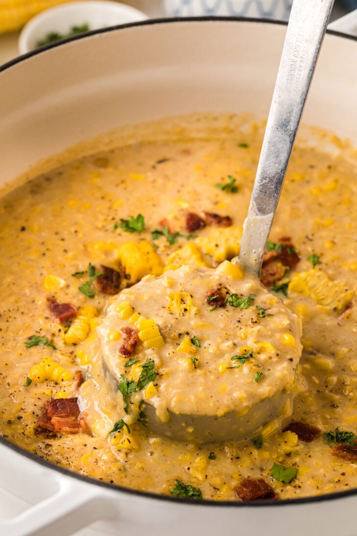Close-up of creamy corn chowder in a white Dutch oven; a ladle lifts a hearty scoop with sweet corn kernels, bacon bits, parsley, and black pepper.