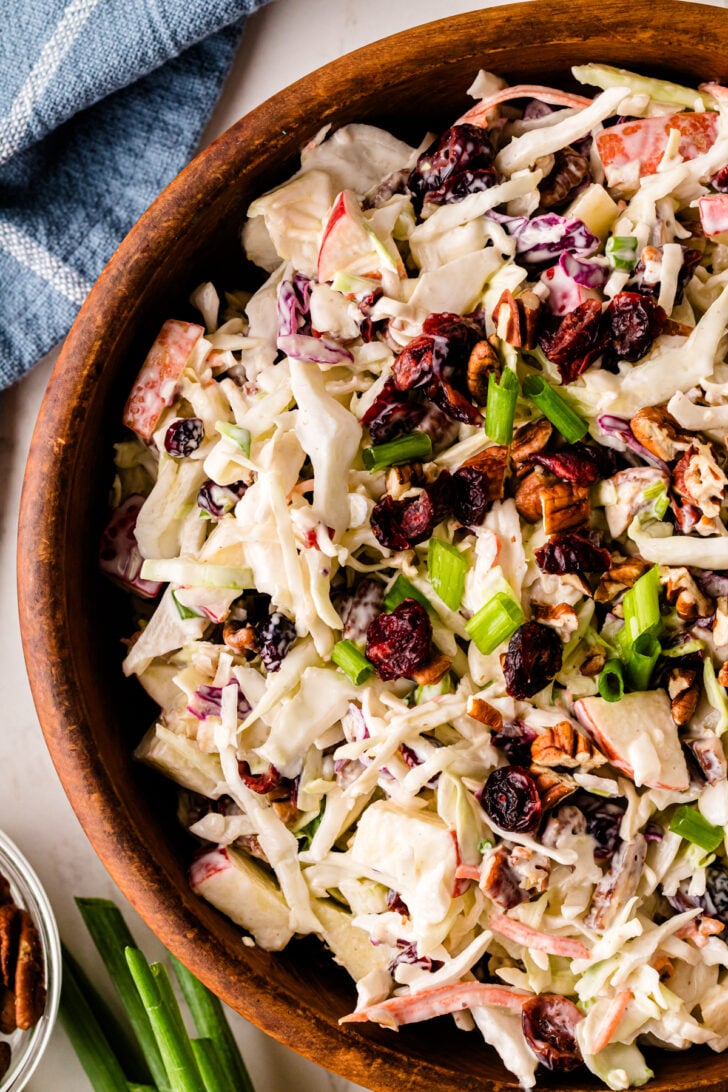 Close-up of creamy coleslaw in a wooden bowl, made with shredded cabbage, apples, dried cranberries, pecans, and green onions.