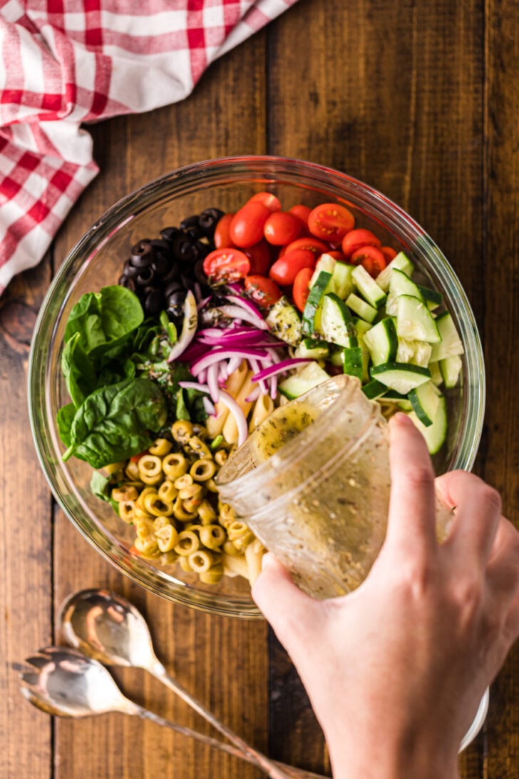 Hand drizzling dressing into glass bowl of Mediterranean Pasta Salad with tomatoes, cucumbers, olives, and pasta
