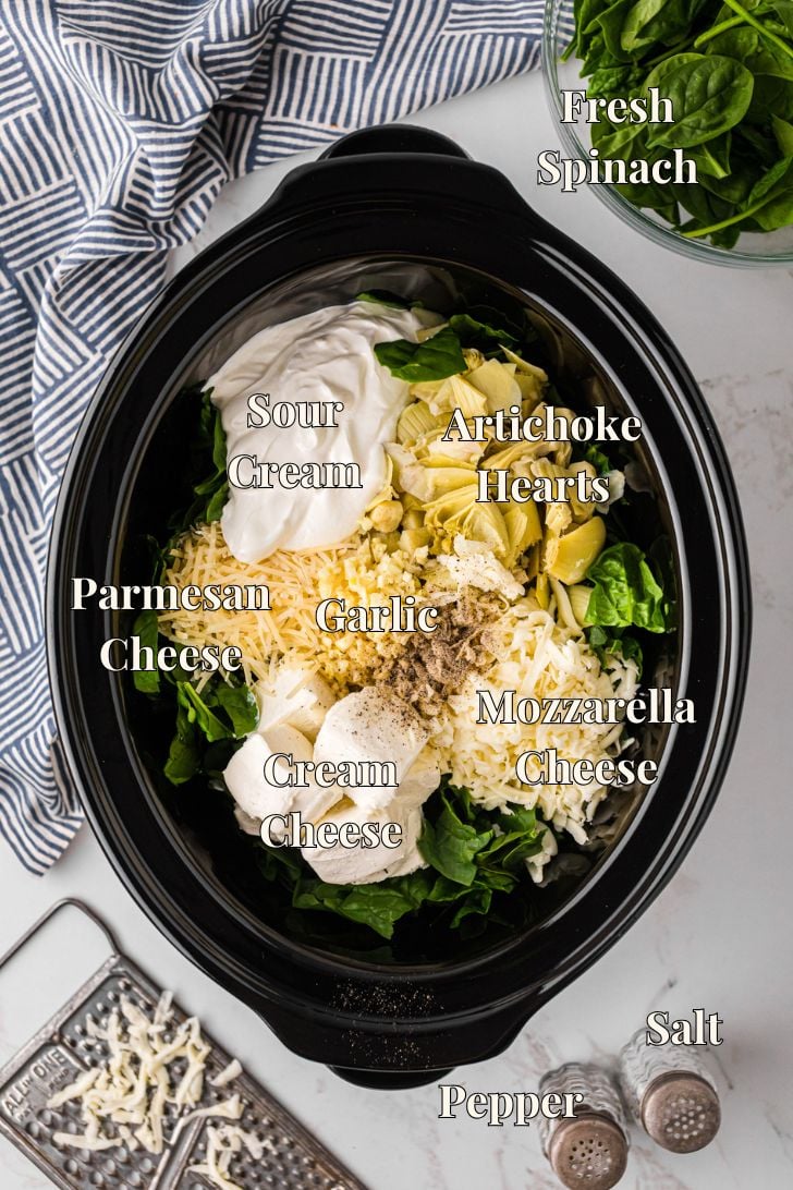 Overhead view of slow cooker filled with spinach artichoke dip ingredients: fresh spinach, artichoke hearts, sour cream, cream cheese, shredded mozzarella, parmesan, garlic, salt, and pepper.