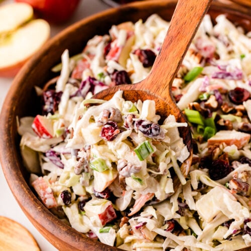 Wooden spoon lifting creamy Cranberry Pecan Slaw from a wooden bowl, topped with shredded cabbage, dried cranberries, pecans, and green onio