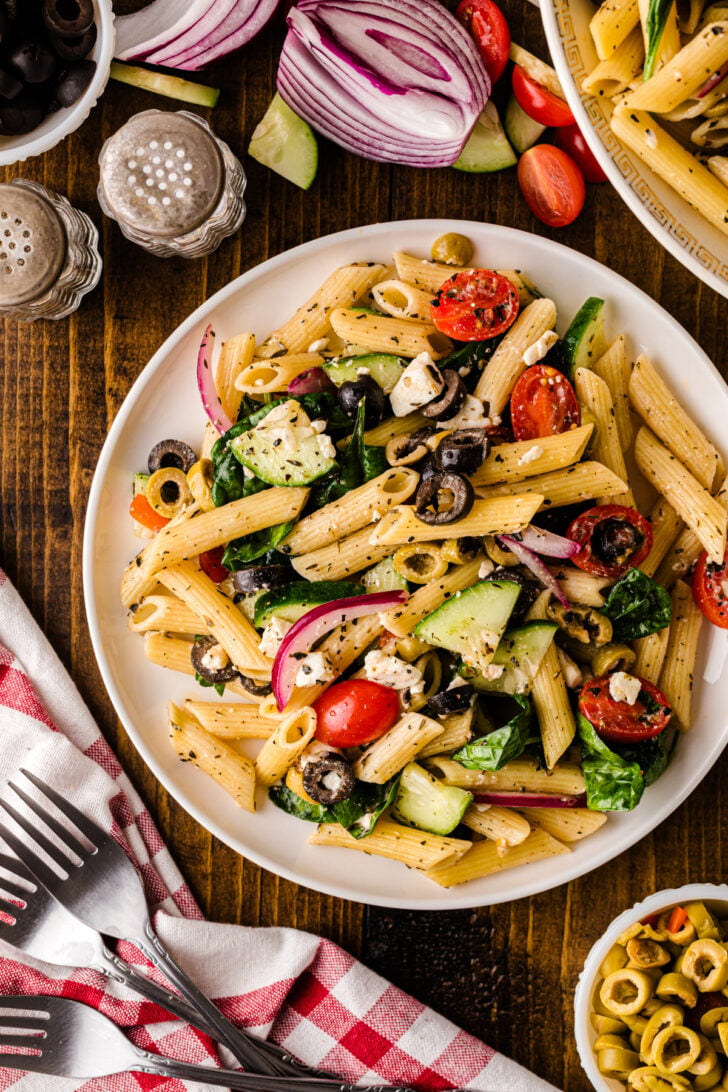 Close-up of Mediterranean pasta salad with penne, cherry tomatoes, cucumbers, spinach, black and green olives, red onion, and feta cheese, tossed with herbs and served in a patterned bowl.
