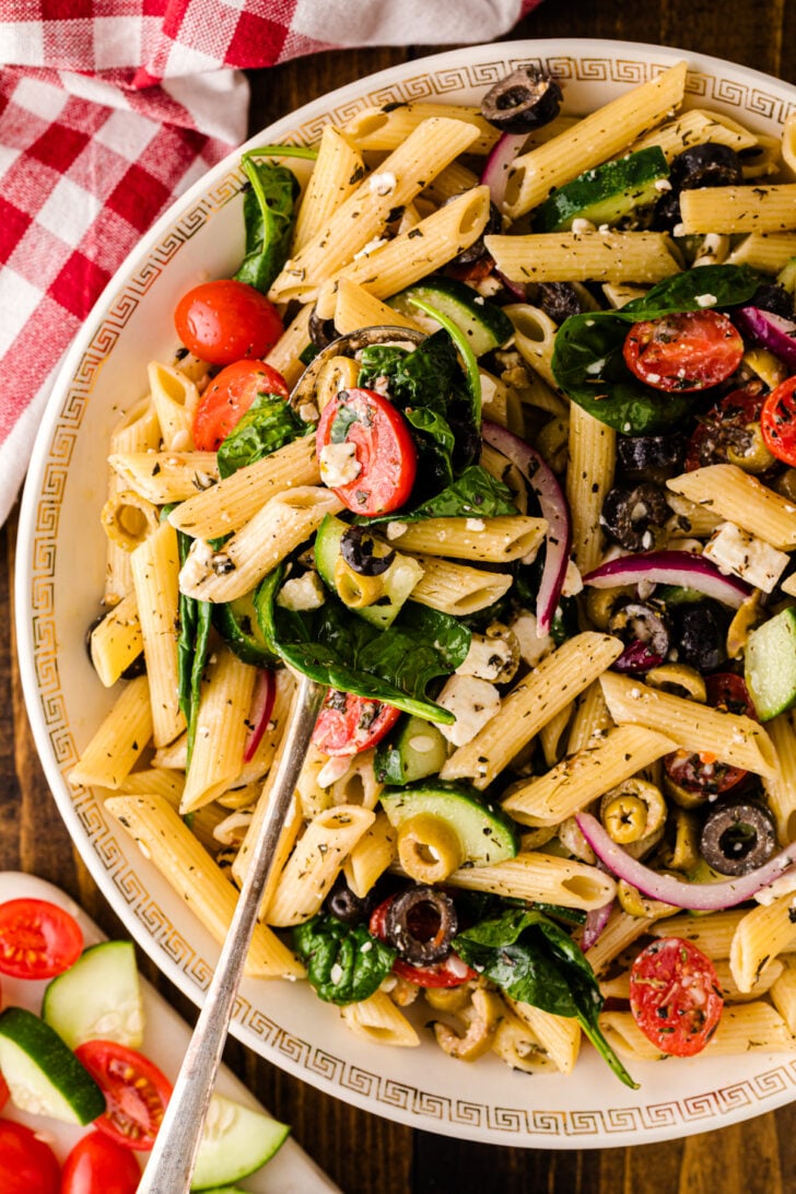 Mediterranean Pasta Salad with penne, tomatoes, cucumbers, olives, spinach, and feta in a white decorative bowl with serving spoon