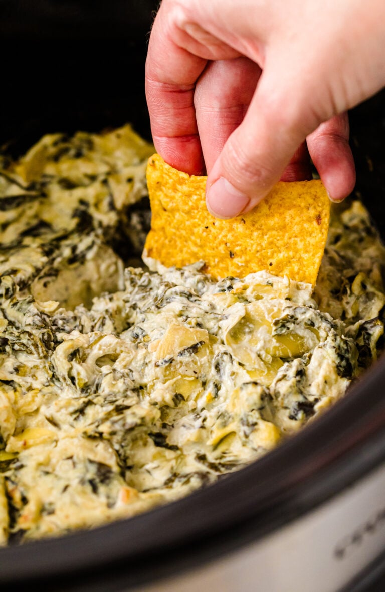 Hand dipping tortilla chip into creamy spinach artichoke dip 4 in dark bowl, herbs visible on surface