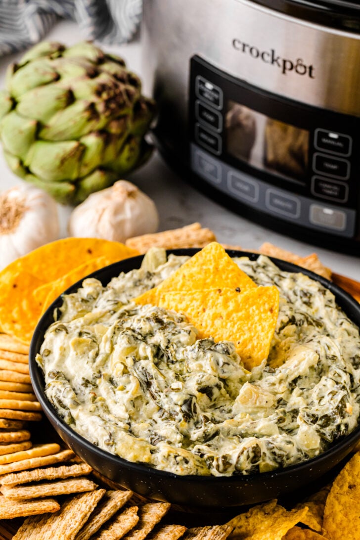 Bowl of creamy spinach artichoke dip served with tortilla chips and crackers, with a slow cooker, garlic, and fresh artichoke in the background.