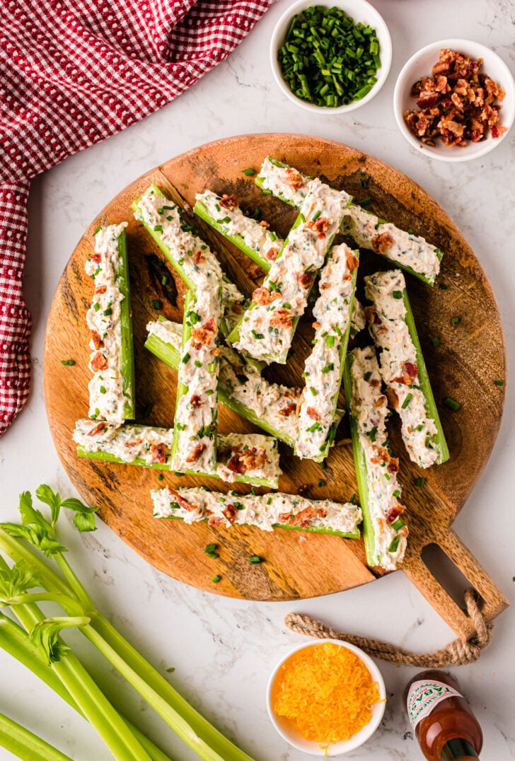 Overhead view of celery sticks stuffed with a creamy cheese mixture, topped with crumbled bacon and chives, arranged on a wooden serving board with bowls of extra toppings nearby.